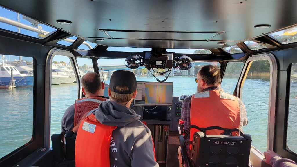 Three people in orange life vests sit in a boat cabin, facing a waterway with a bridge, other boats, and a navigation screen showing data.