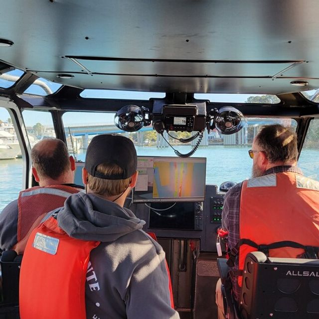 Three people in orange life vests sit in a boat cabin, facing a waterway with a bridge, other boats, and a navigation screen showing data.
