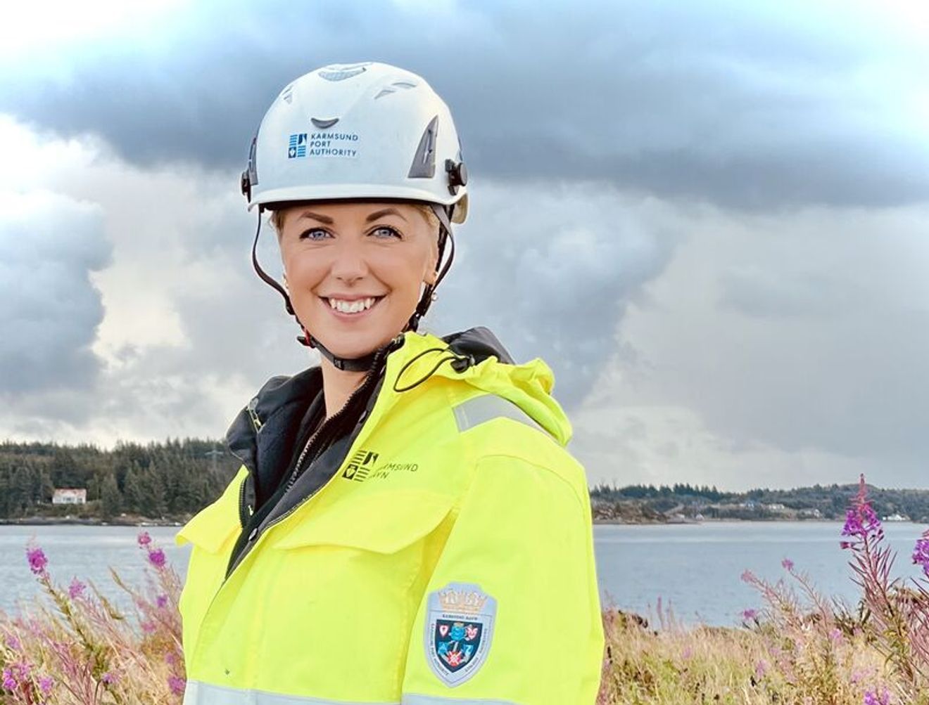 A smiling woman in a white hard hat and a neon yellow and black high-visibility jacket stands in a field of purple wildflowers by the water.