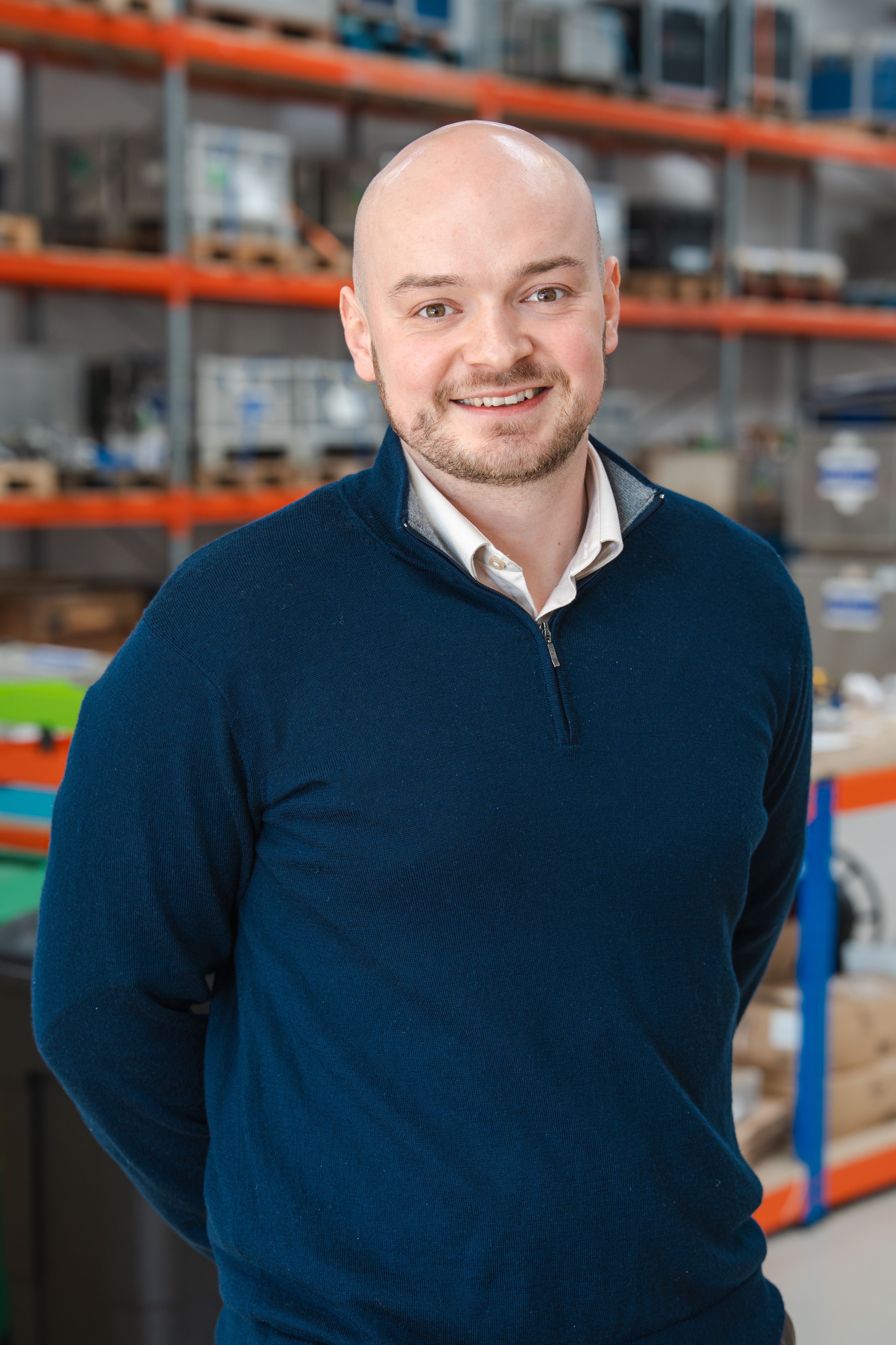 A smiling bald man with a beard wears a blue quarter-zip sweater, standing in a warehouse.