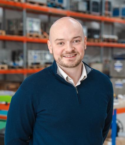 A smiling bald man with a beard wears a blue quarter-zip sweater, standing in a warehouse.