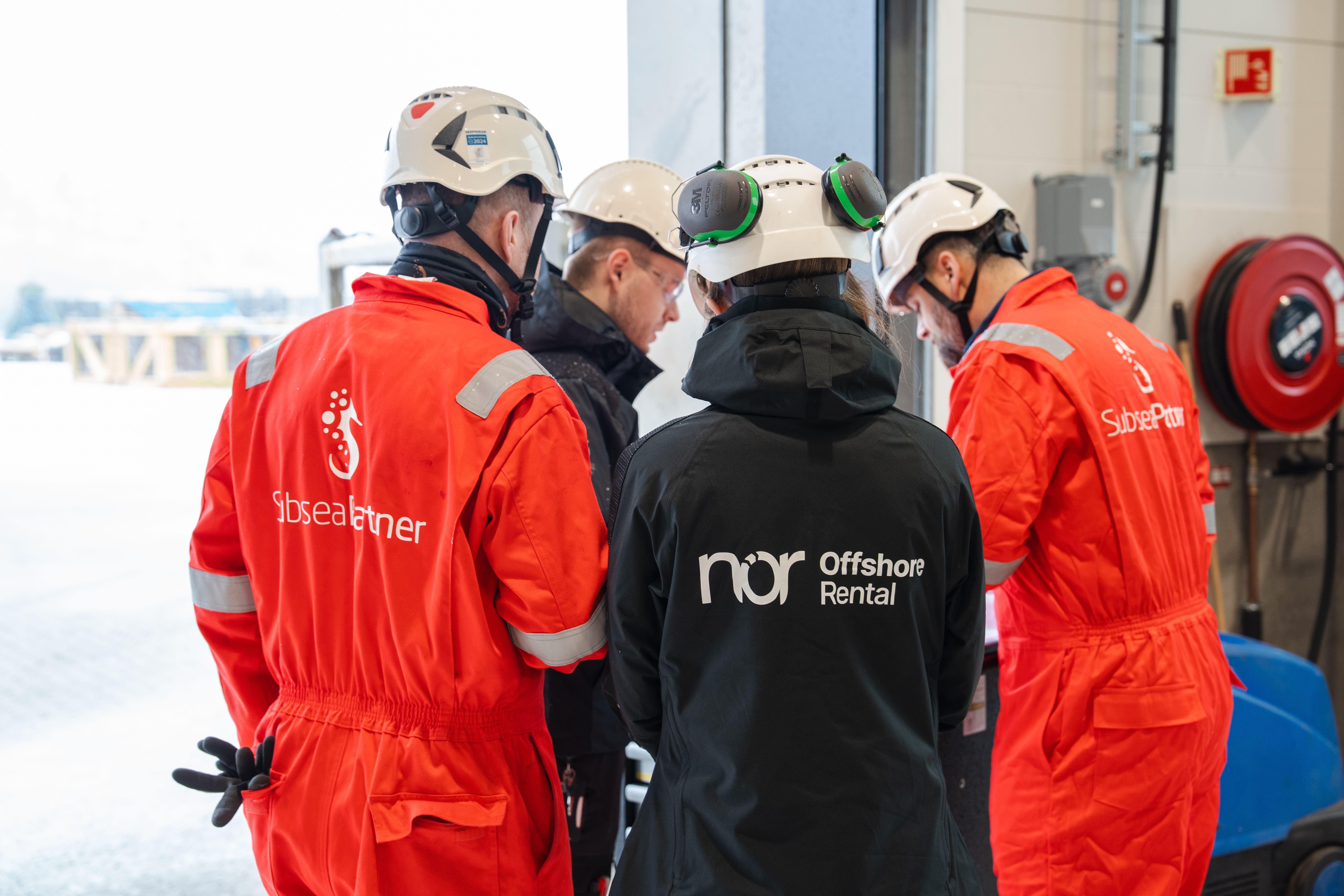 Four people in hard hats and industrial workwear, seen from behind, gathered and observing something.