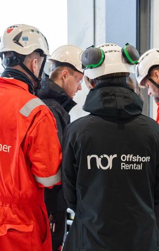 Four people in hard hats and industrial workwear, seen from behind, gathered and observing something.