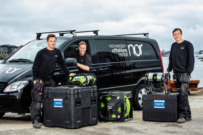 Three men pose with a black "Norwegian Offshore Rental" van and marine ROV equipment by the water.