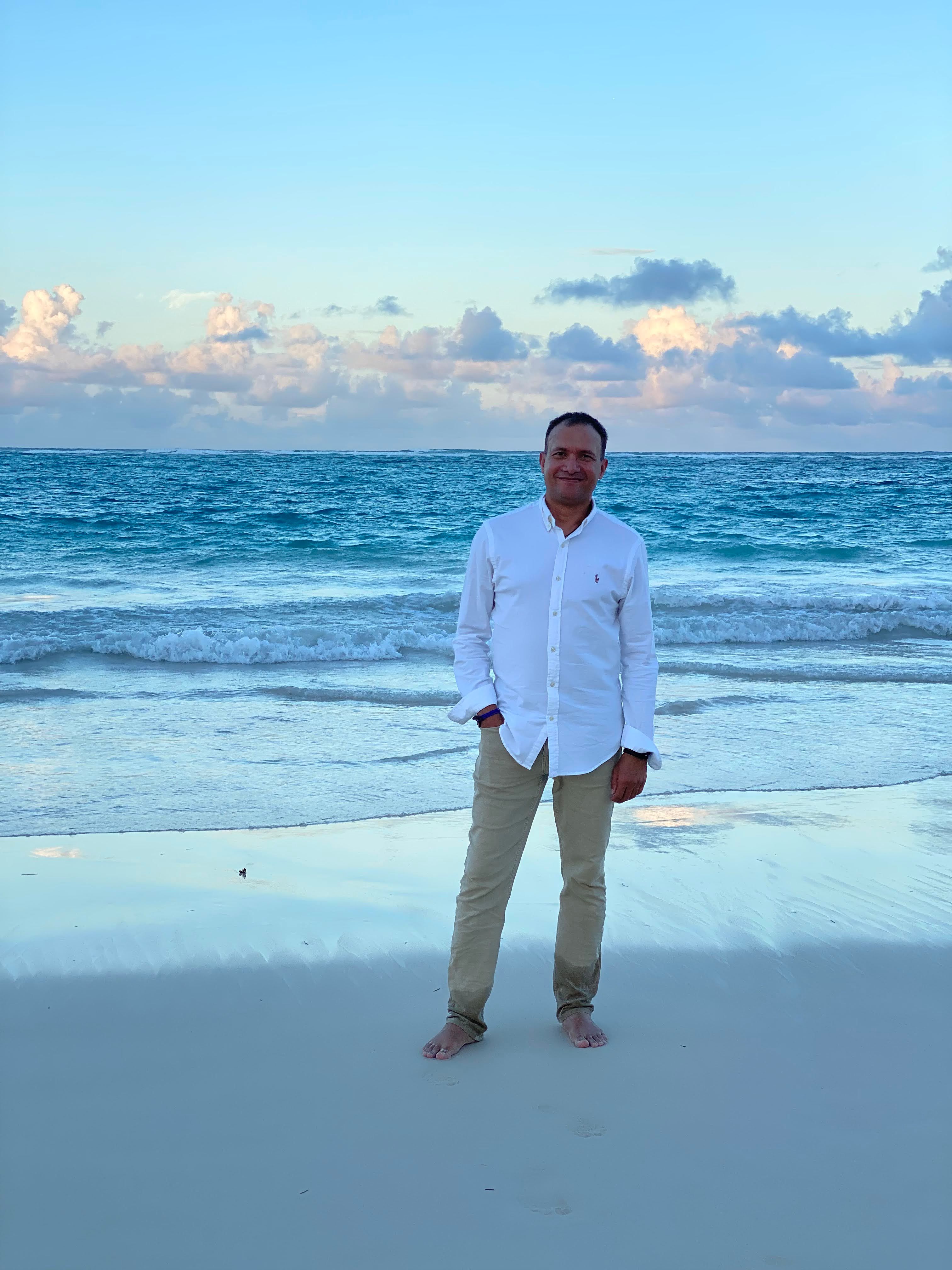 A smiling man in a white shirt and khaki pants stands barefoot on a beach with the ocean and a cloudy sky in the background.