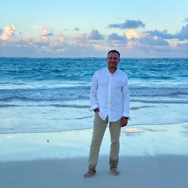 A smiling man in a white shirt and khaki pants stands barefoot on a beach with the ocean and a cloudy sky in the background.