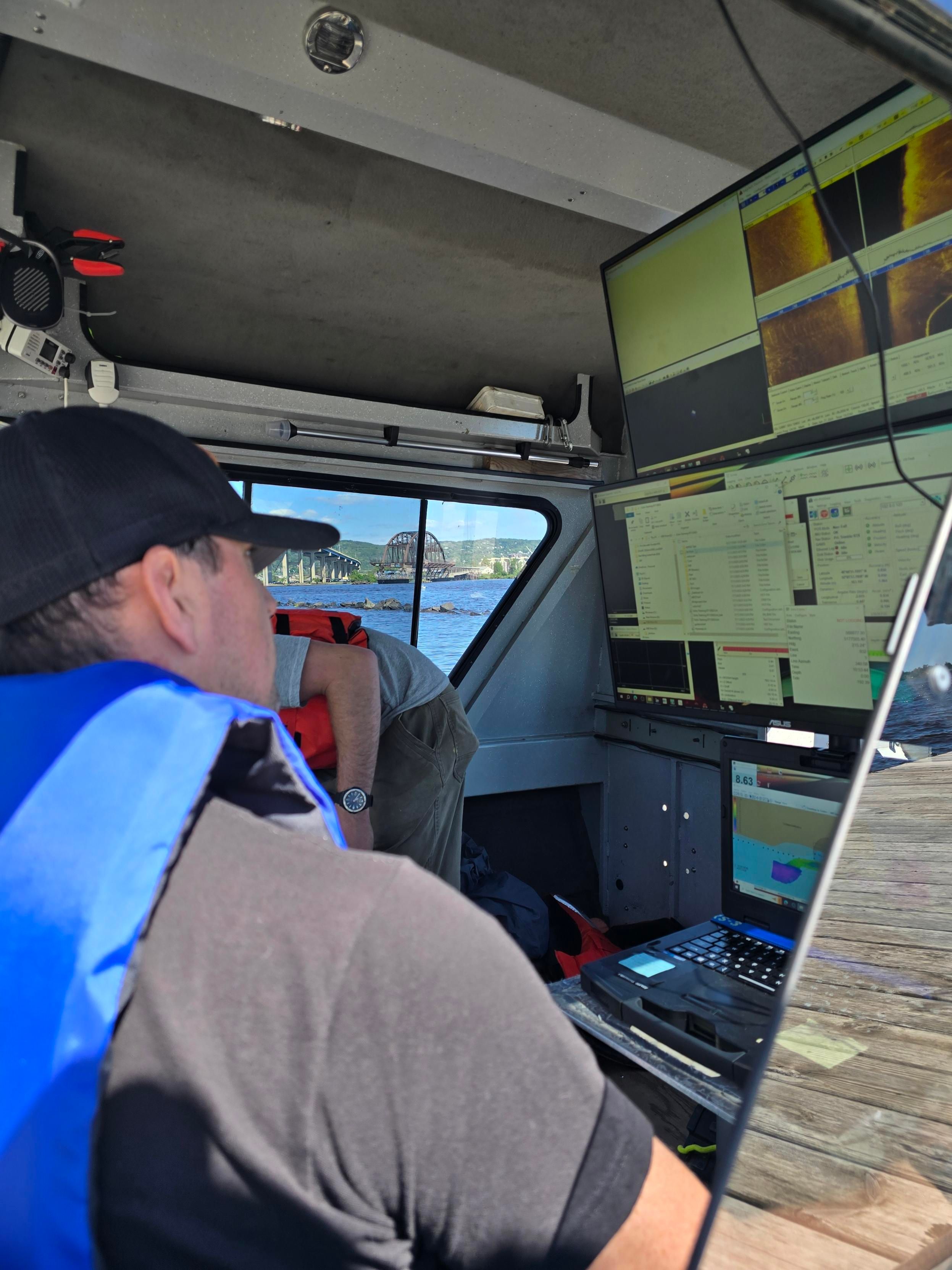 A person in a life vest inside a boat monitors sonar and mapping data on multiple screens, with a bridge and city outside.