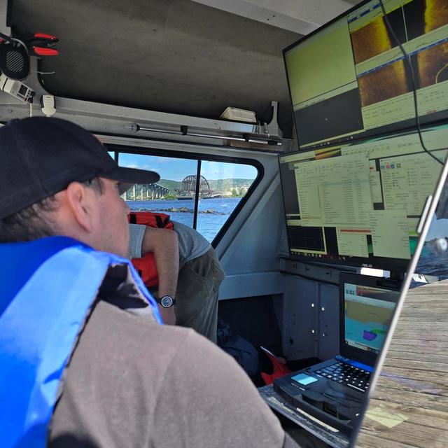 A person in a life vest inside a boat monitors sonar and mapping data on multiple screens, with a bridge and city outside.