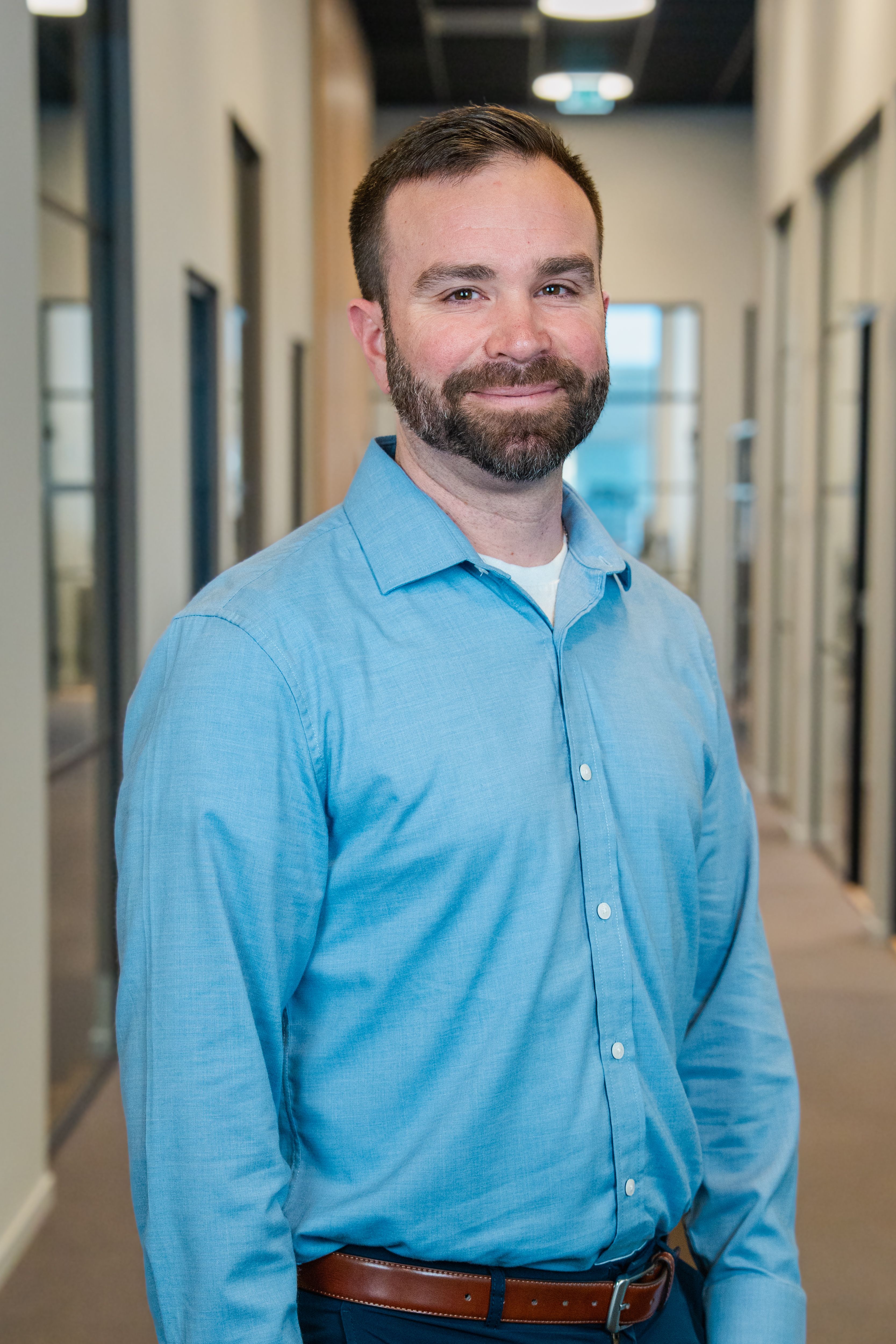 A smiling man with a beard wearing a blue shirt stands in an office hallway.