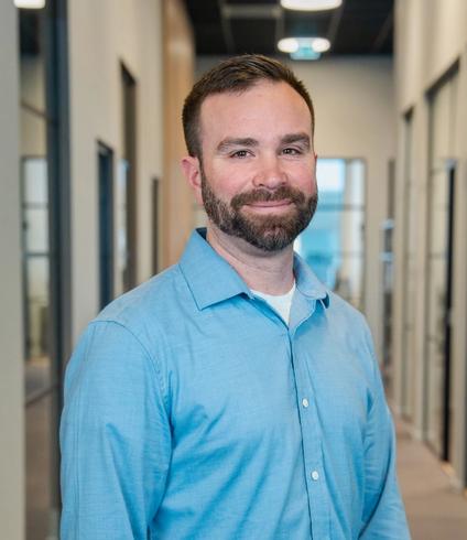 A smiling man with a beard wearing a blue shirt stands in an office hallway.