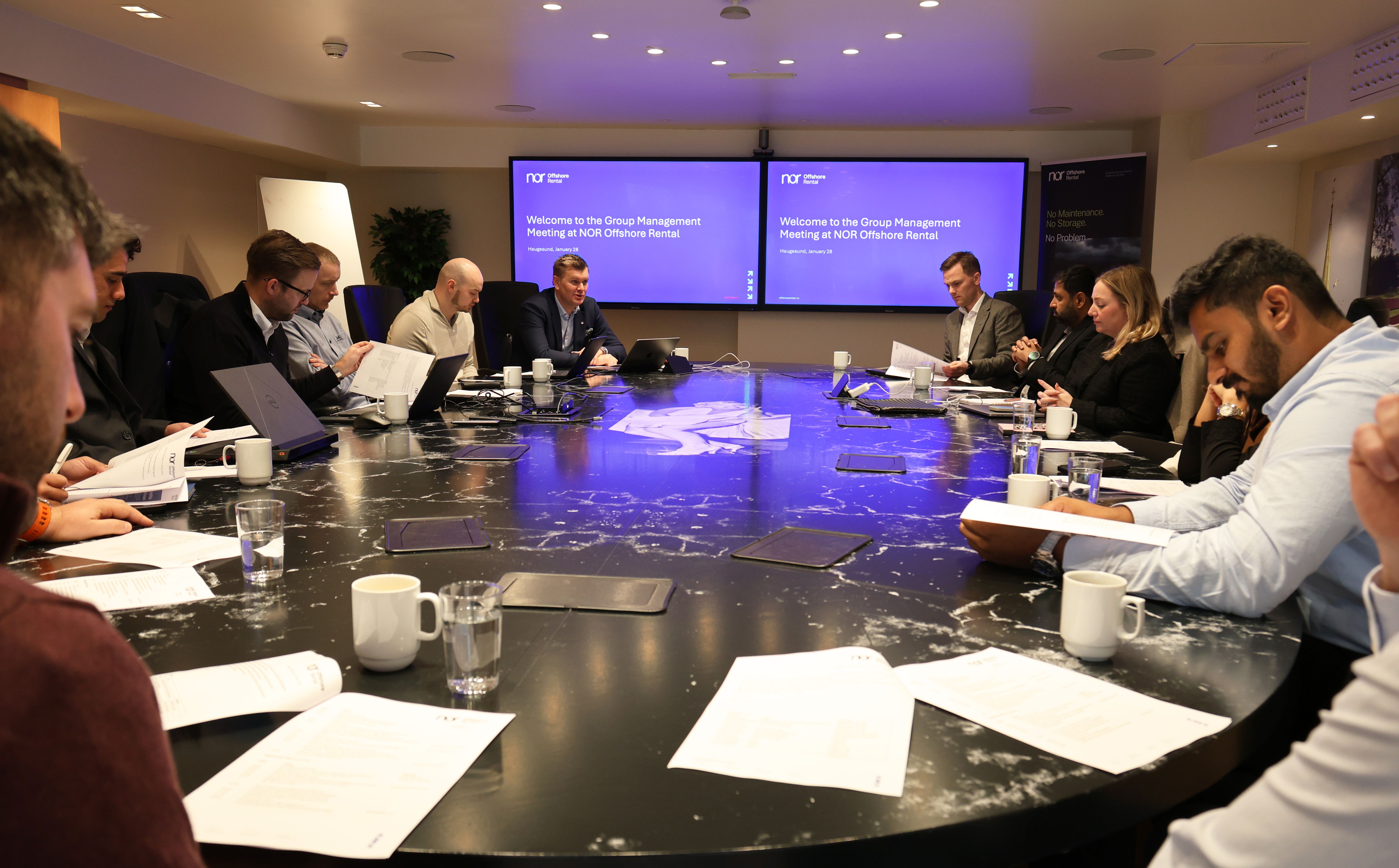 A group of professionals in a conference room seated around a table with laptops and papers, with two screens displaying "Welcome to the Group Management Meeting at NOR Offshore Rental."