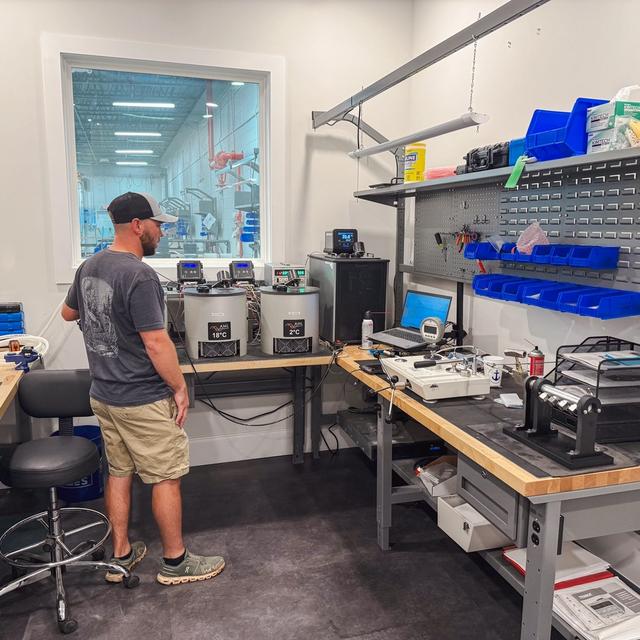 A man stands in a lab, looking through a window into a factory, with scientific equipment and tools on surrounding workbenches.