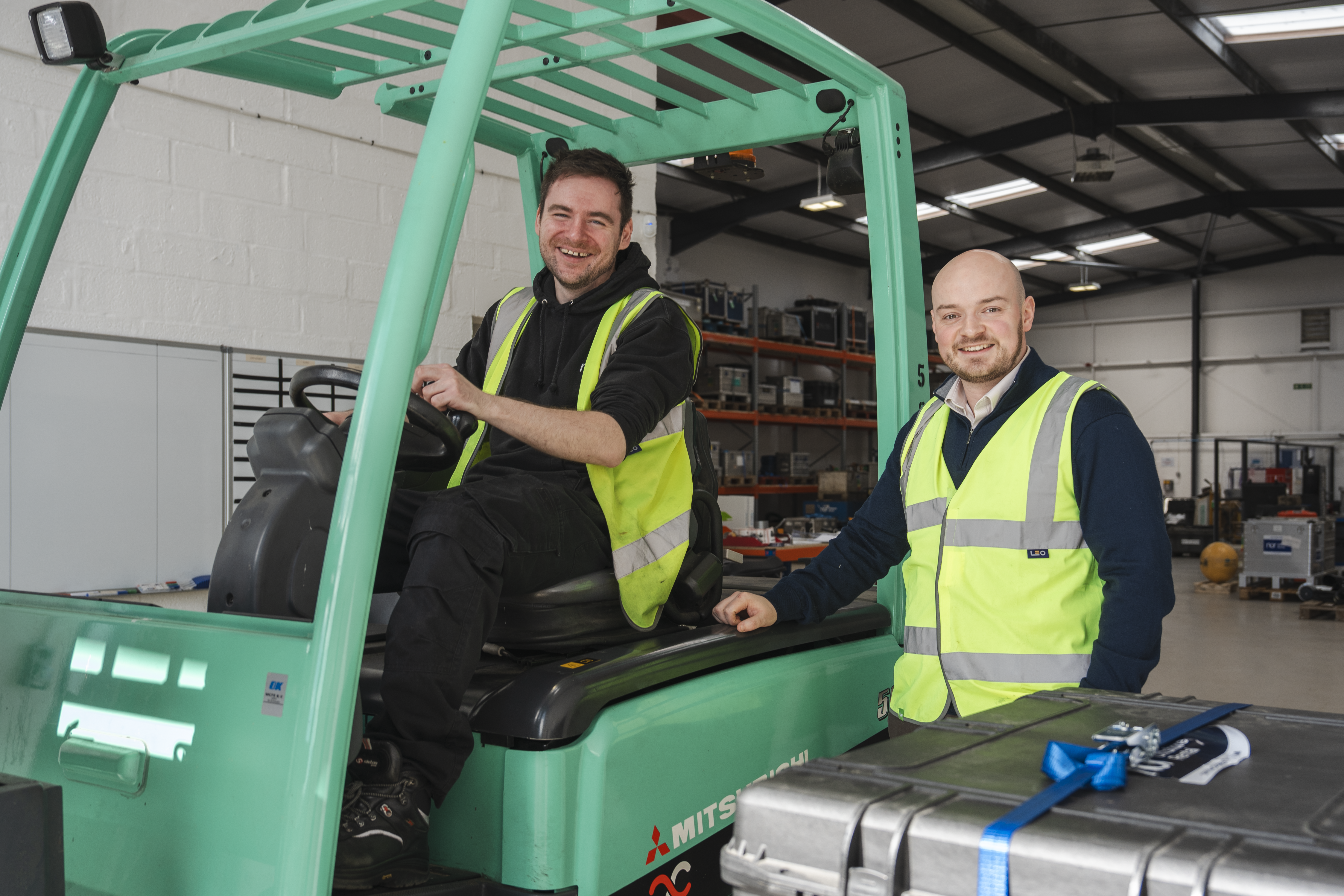 Two smiling men in safety vests with a forklift and a silver case in a warehouse.