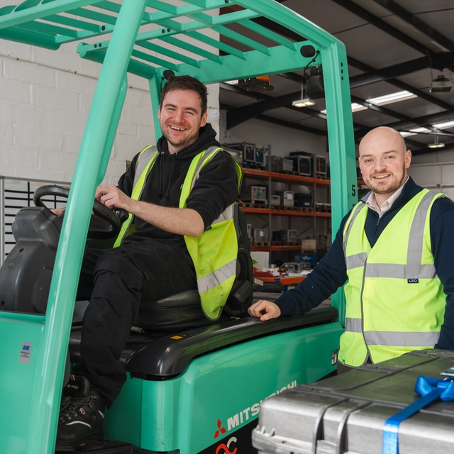 Two smiling men in safety vests with a forklift and a silver case in a warehouse.