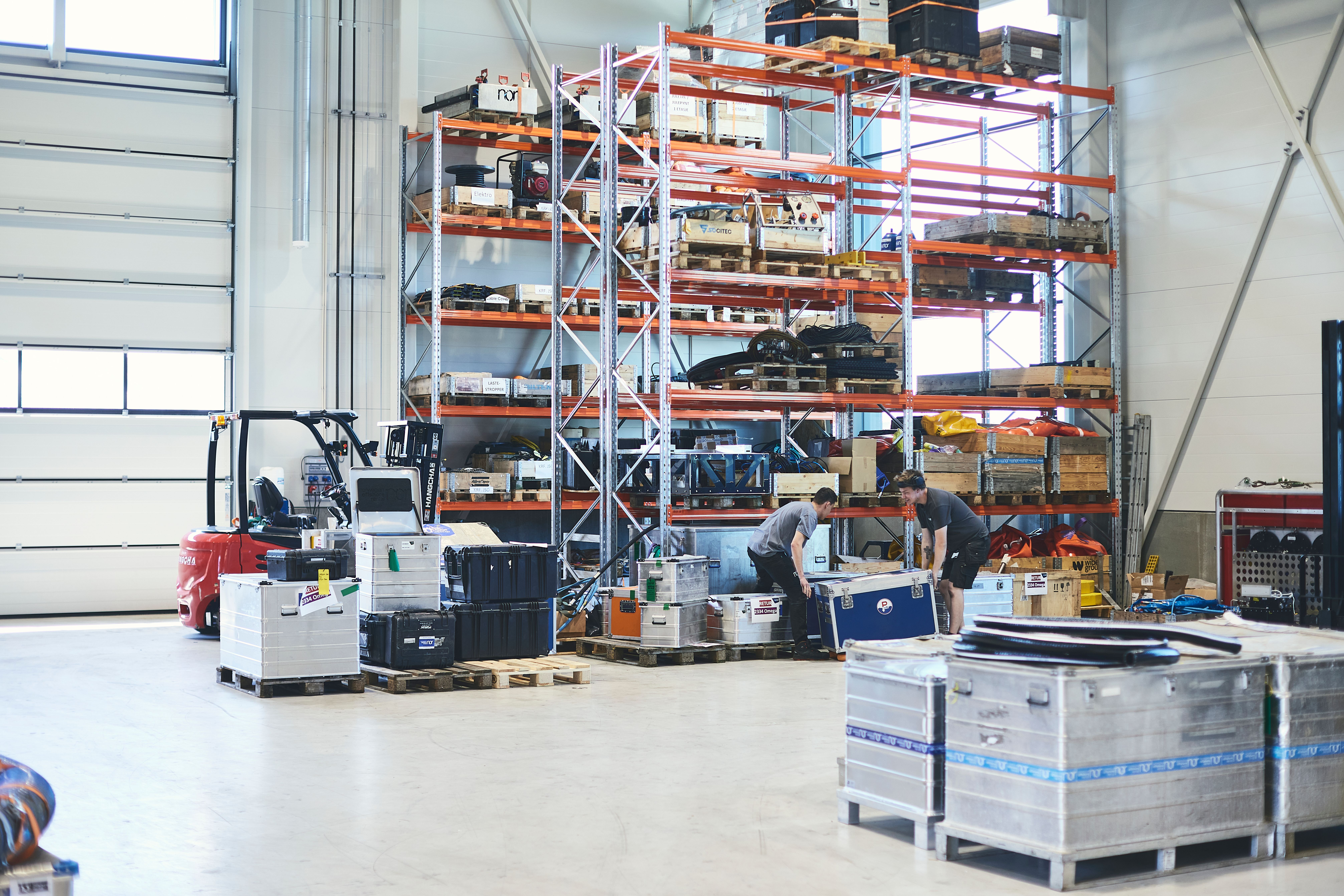Warehouse interior with tall storage racks, a forklift, and two people moving a large box.