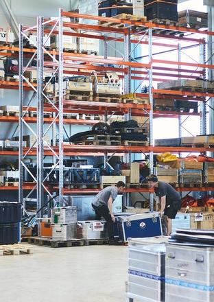Warehouse interior with tall storage racks, a forklift, and two people moving a large box.