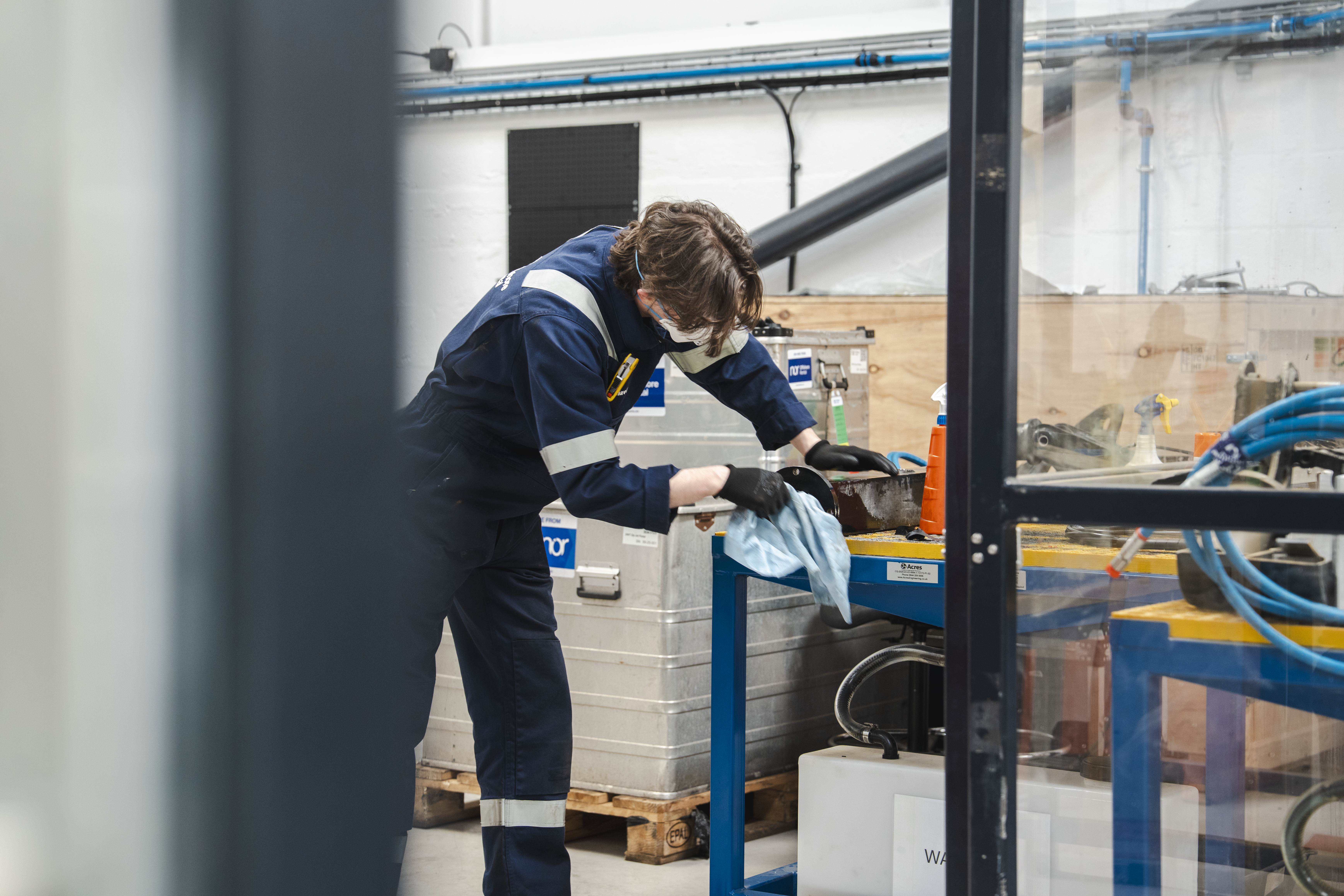 A person in blue overalls and safety glasses wiping an object on a workbench in a workshop.