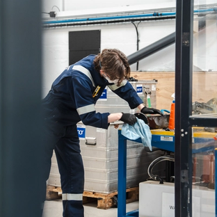 A person in blue overalls and safety glasses wiping an object on a workbench in a workshop.