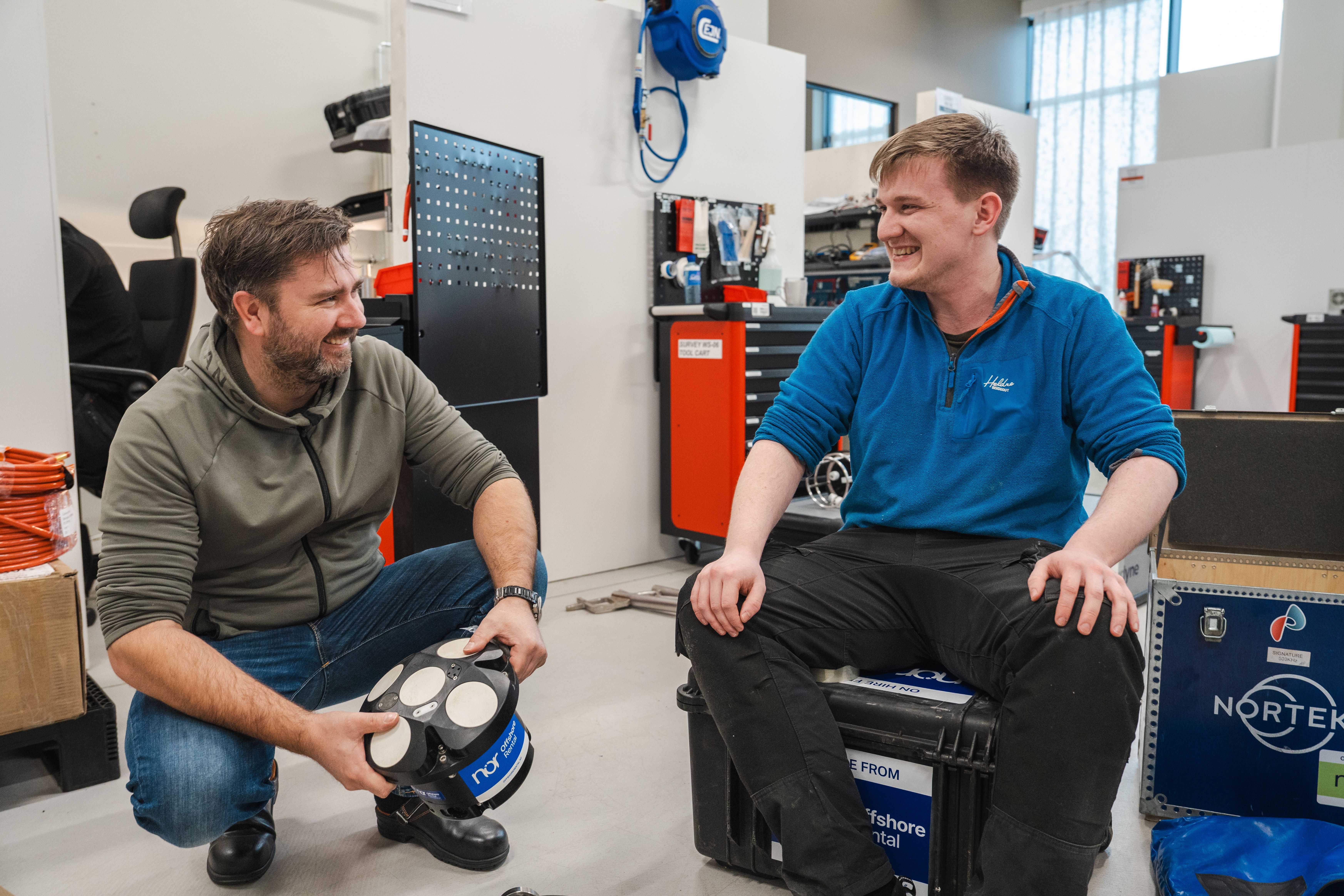 Two NOR employees smiling and talking in a workshop, surrounded by offshore rental equipment and tools.