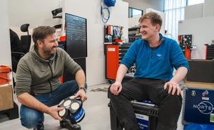 Two men in a workshop, one crouching and holding a Nortek multi-sensor device while smiling at the other man sitting on a case.
