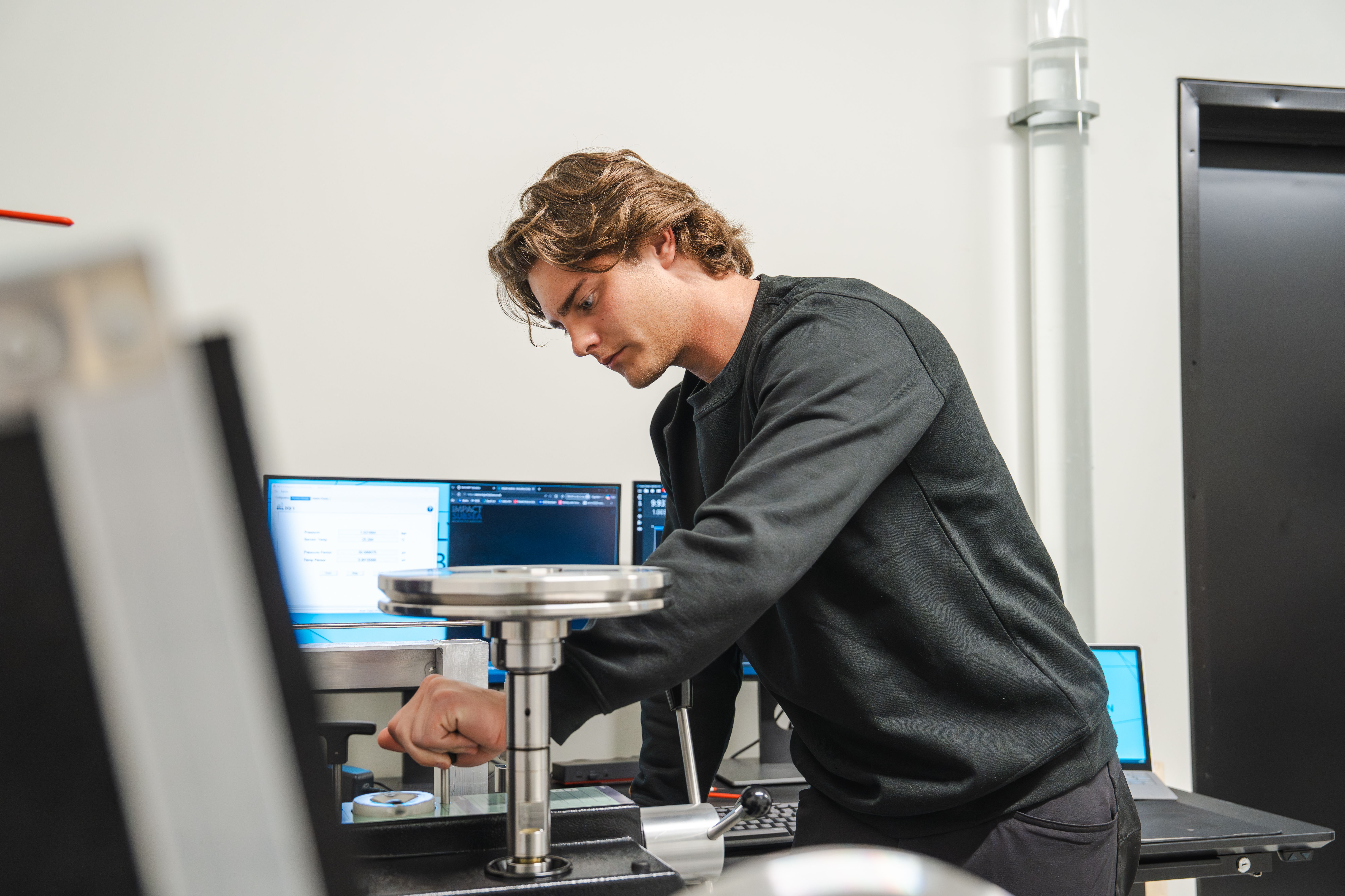 A young man in a black shirt operates lab equipment, with computer monitors in the background.
