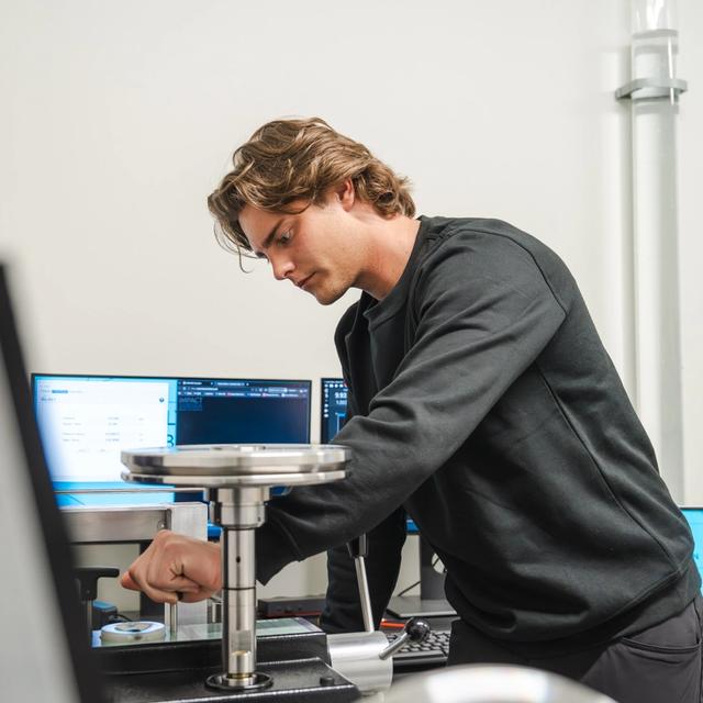 A young man in a black shirt operates lab equipment, with computer monitors in the background.