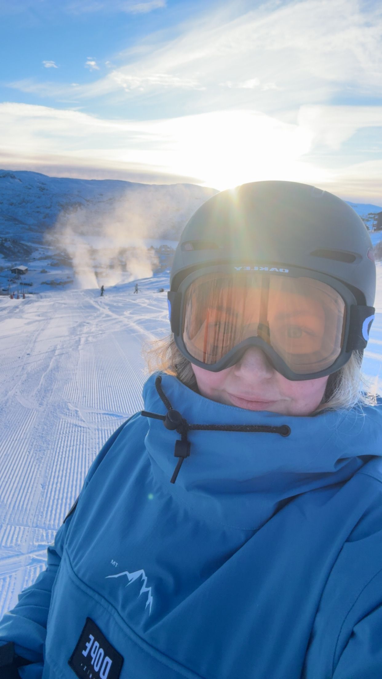 A person in a gray ski helmet and orange reflective goggles stands on a sunny, groomed ski slope, with bright sun glare reflecting off the goggles and snowy mountains and steam in the background.