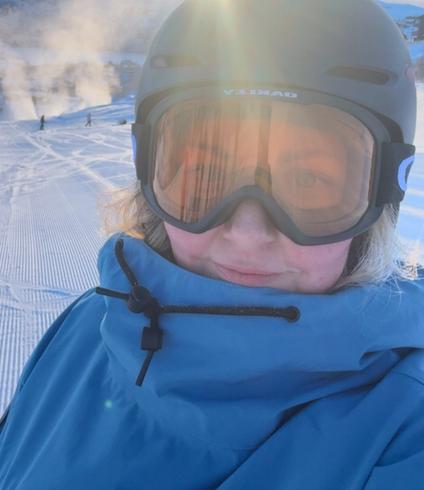 A person in a gray ski helmet and orange reflective goggles stands on a sunny, groomed ski slope, with bright sun glare reflecting off the goggles and snowy mountains and steam in the background.