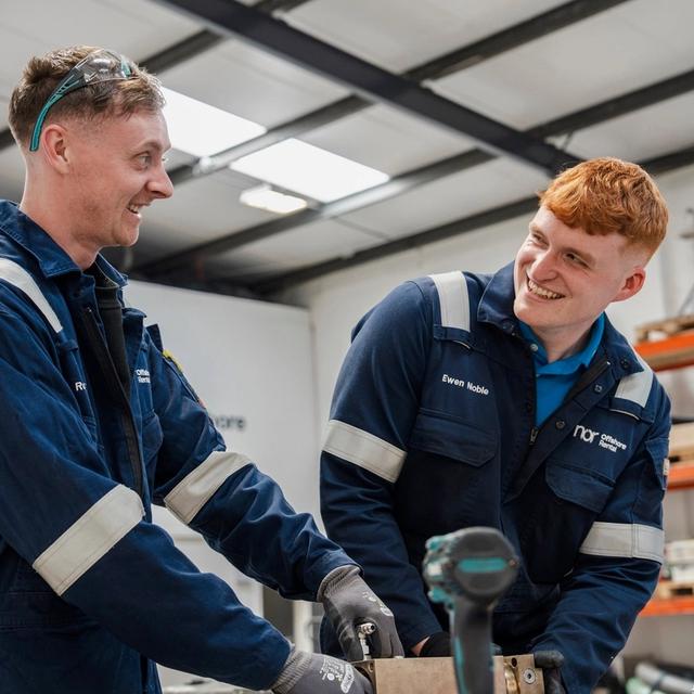 Two smiling workers in blue overalls conversing in a workshop.