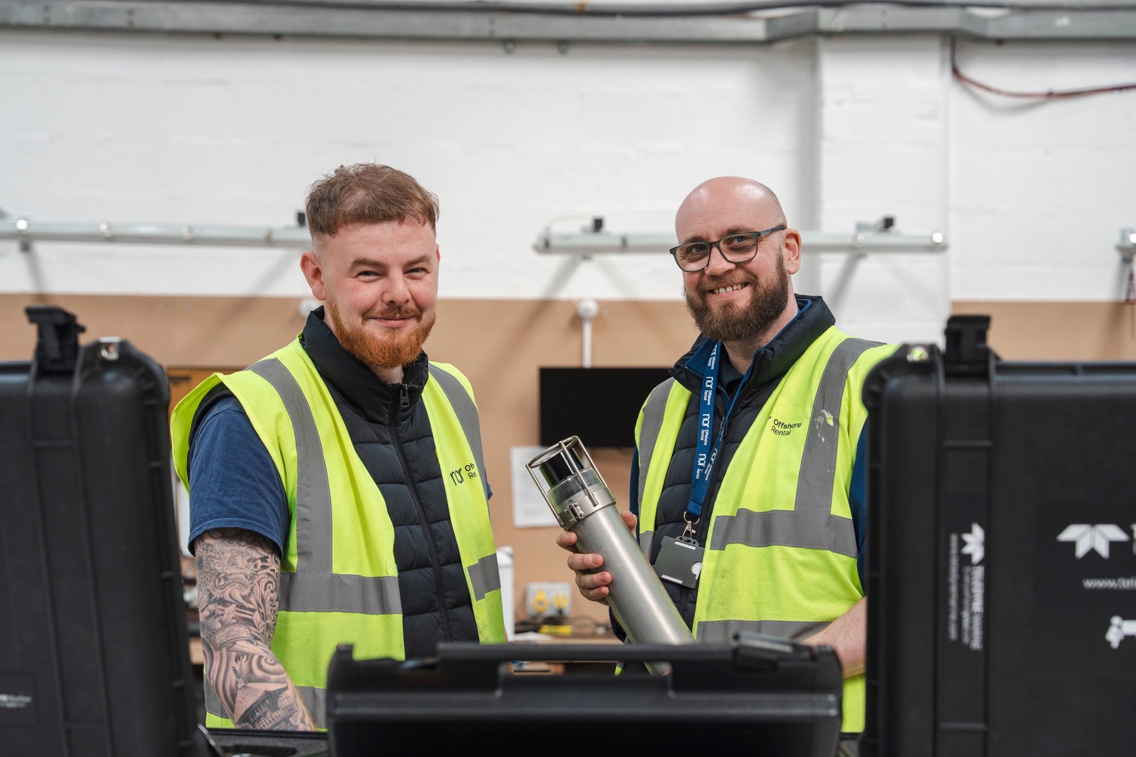 Two smiling men in high-visibility vests, one holding a piece of equipment, in a workshop.