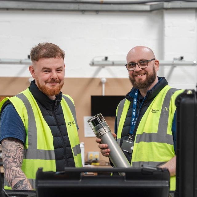 Two smiling men in high-visibility vests, one holding a piece of equipment, in a workshop.