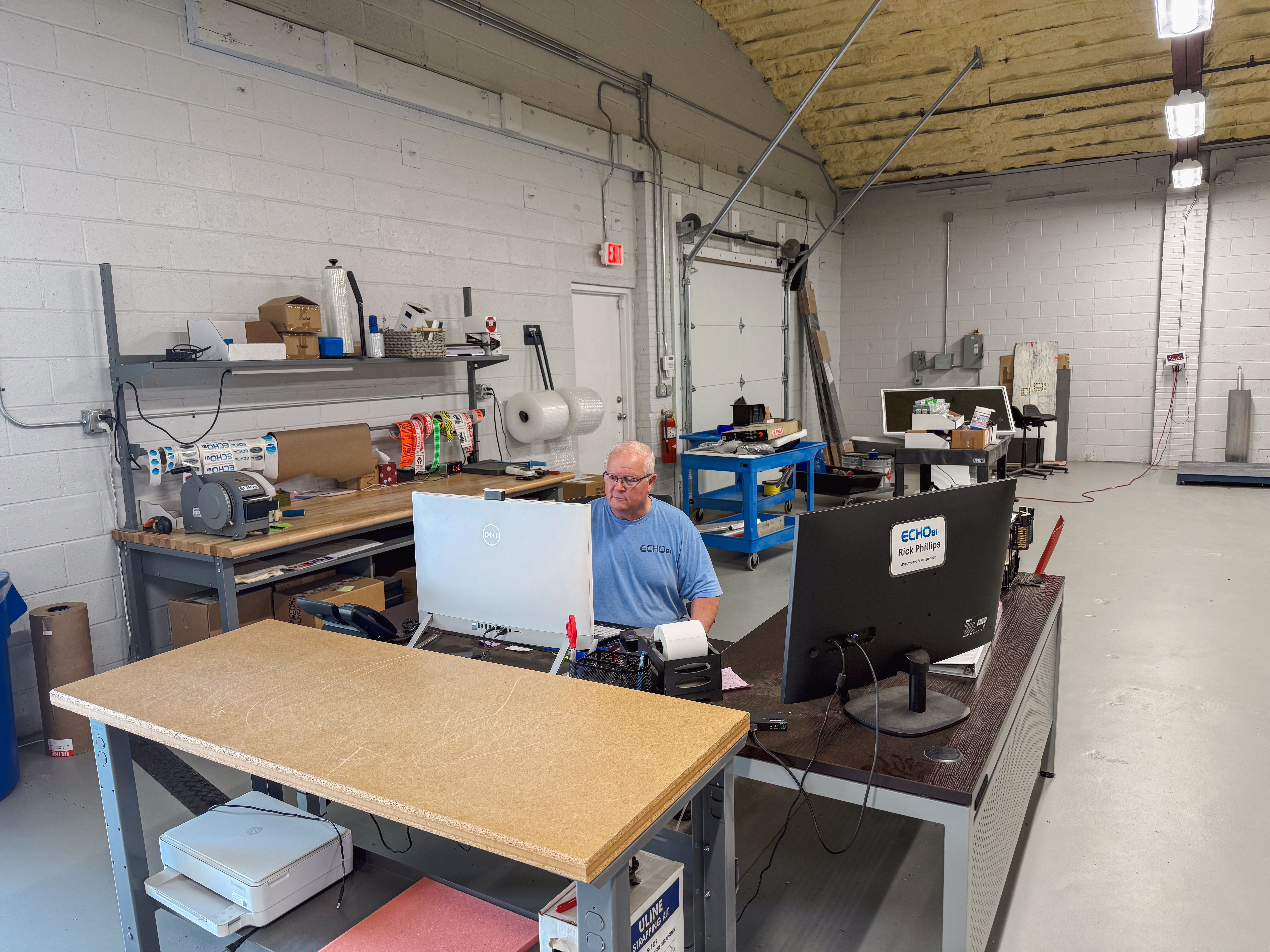 A man wearing a blue shirt and glasses works at a desk with two monitors in a warehouse office.