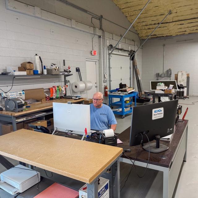 A man wearing a blue shirt and glasses works at a desk with two monitors in a warehouse office.