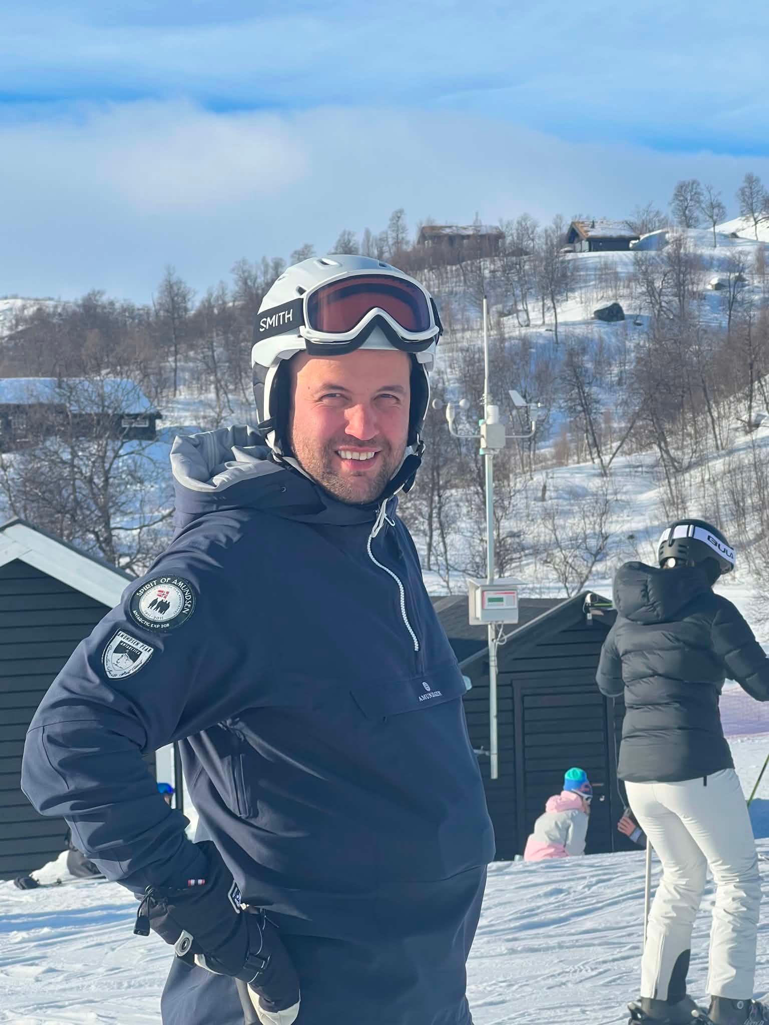 A man in a white helmet and dark blue jacket smiles on a snowy mountain with buildings and trees in the background.