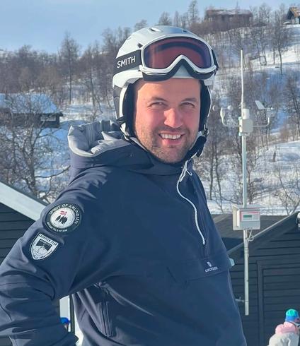 A man in a white helmet and dark blue jacket smiles on a snowy mountain with buildings and trees in the background.