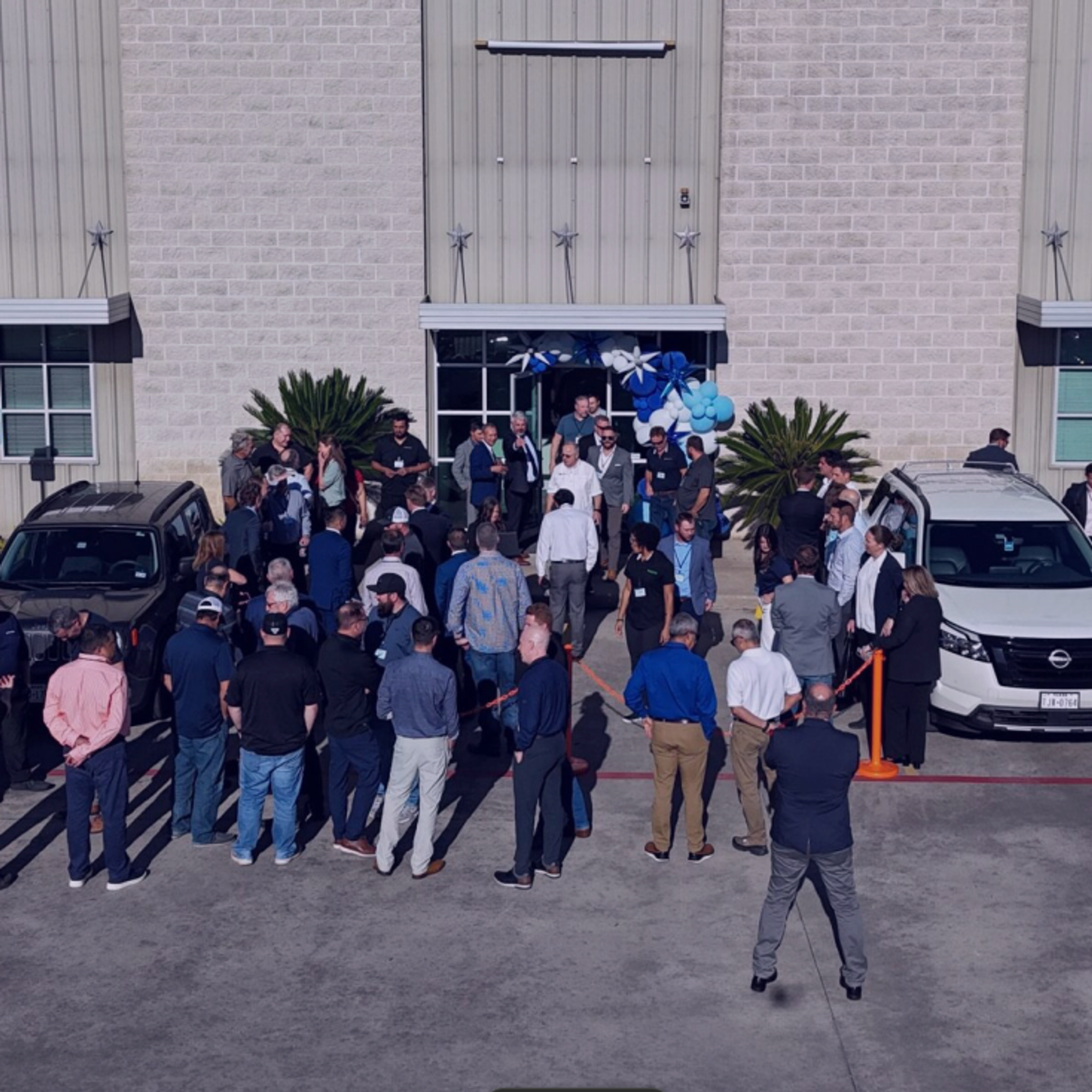 A crowd of people gathered outside a commercial building with cars and blue and white decorations.