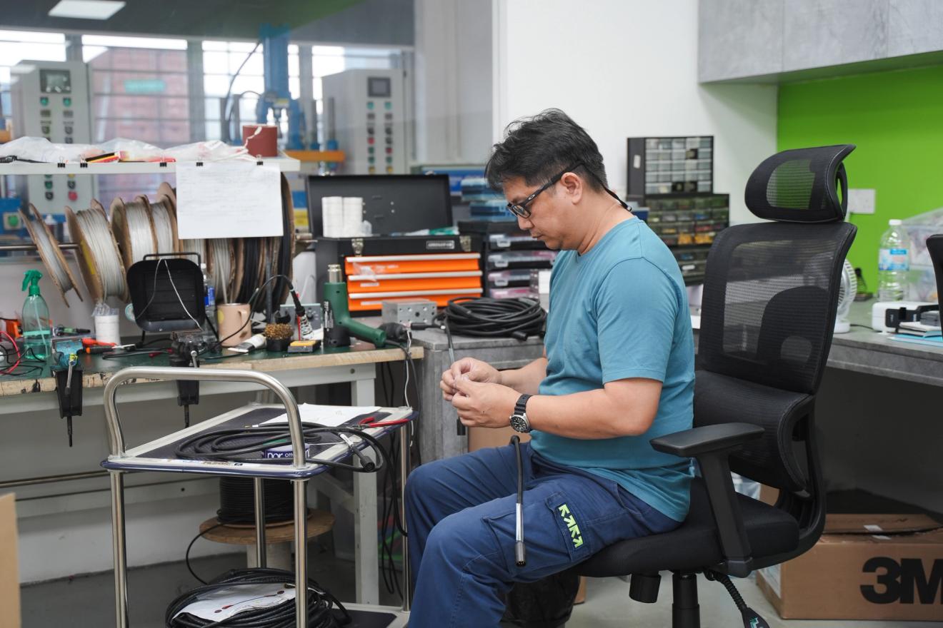 A man in a blue t-shirt sits at a workbench, focused on assembling or repairing a cable.