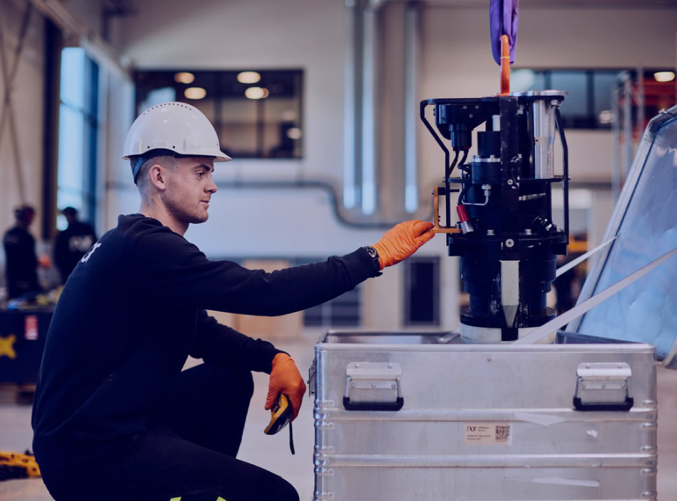 A man in a hard hat and gloves adjusts machinery being lowered into a metal crate in a warehouse.