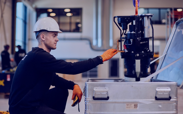 A man in a hard hat and gloves adjusts machinery being lowered into a metal crate in a warehouse.