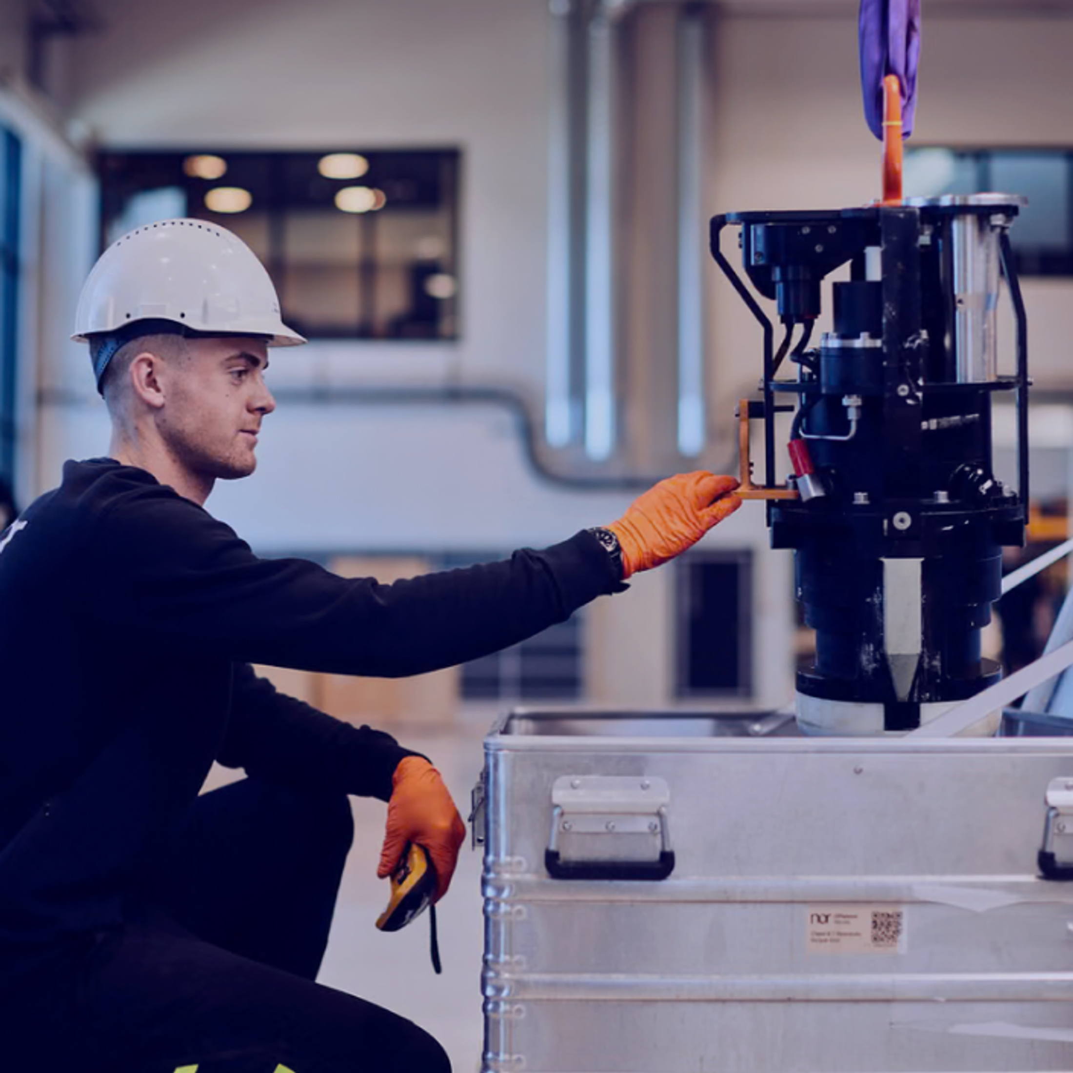 A man in a hard hat and gloves adjusts machinery being lowered into a metal crate in a warehouse.