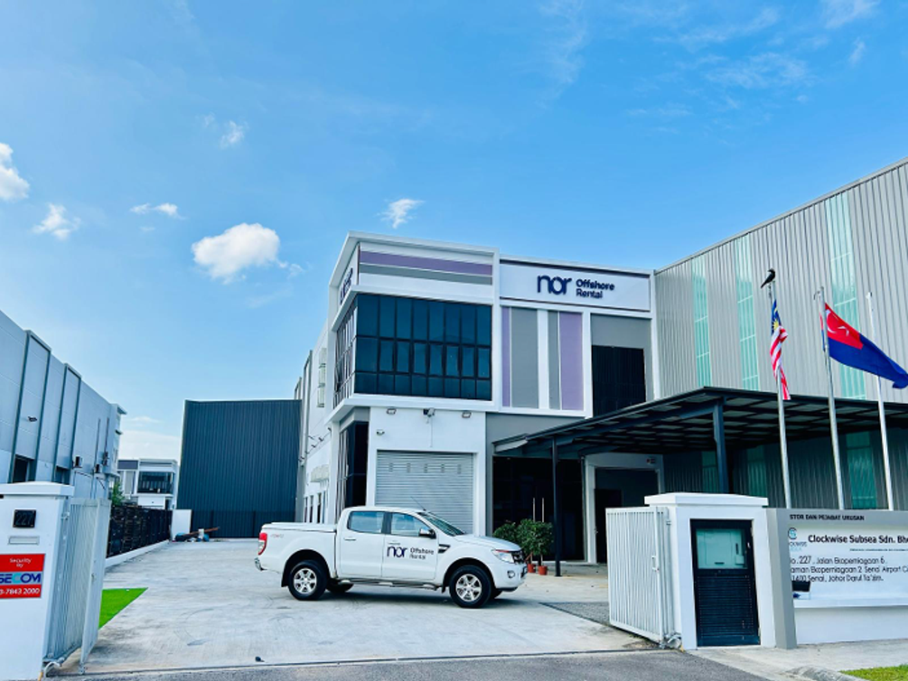 Exterior view of a modern commercial building with "nor Offshore Rental" signage, a white pickup truck, and two flags flying under a blue sky.