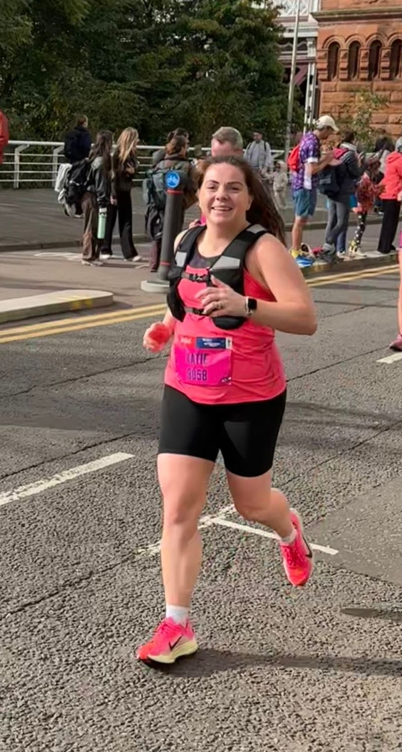 A smiling woman in a pink top, black shorts, and a running bib that reads "KATIE 8058" runs a race.