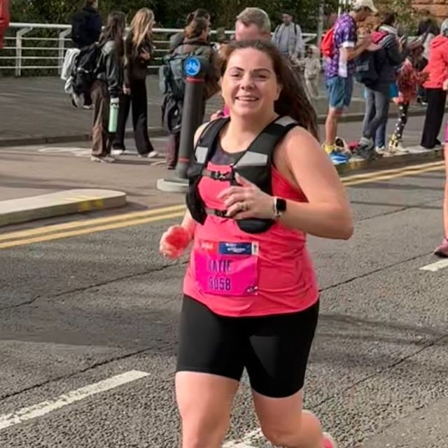 A smiling woman in a pink top, black shorts, and a running bib that reads "KATIE 8058" runs a race.