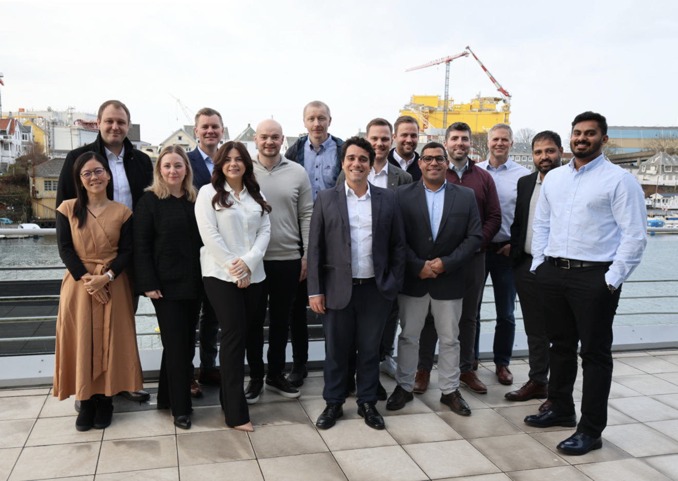 A group of 15 diverse professionals smiling outdoors on a waterfront patio with city buildings and a crane in the background.