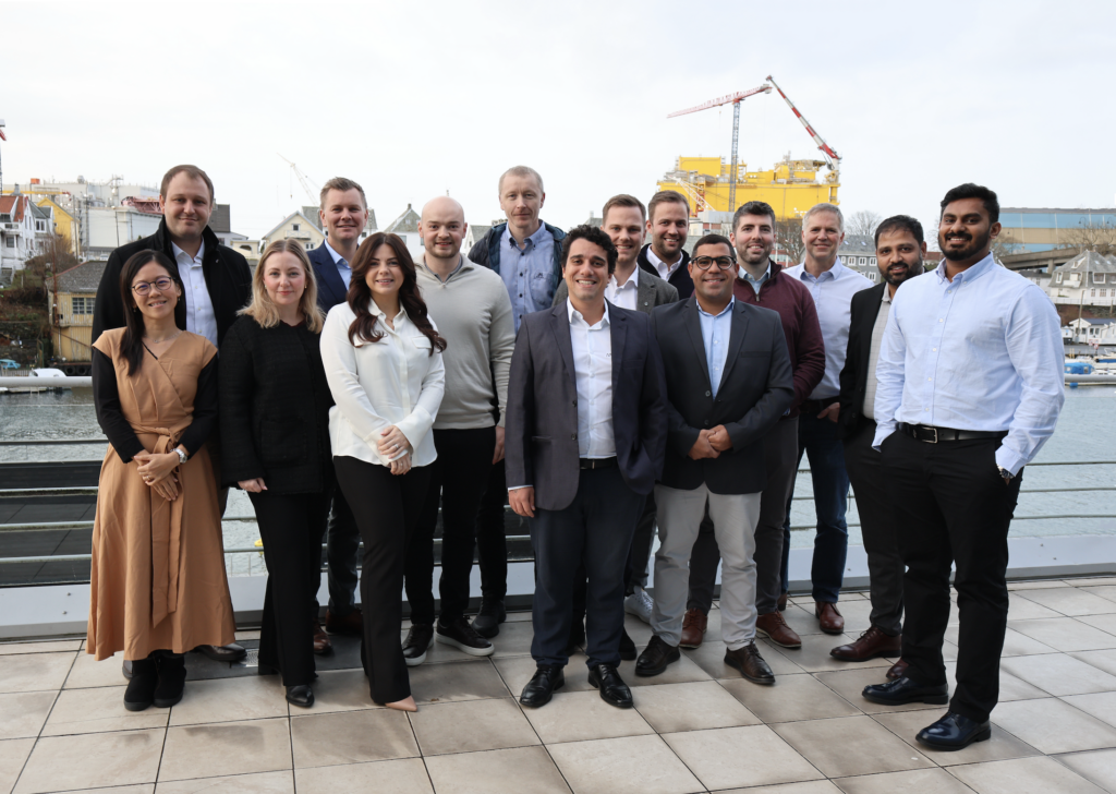 A group of 15 diverse professionals smiling outdoors on a waterfront patio with city buildings and a crane in the background.