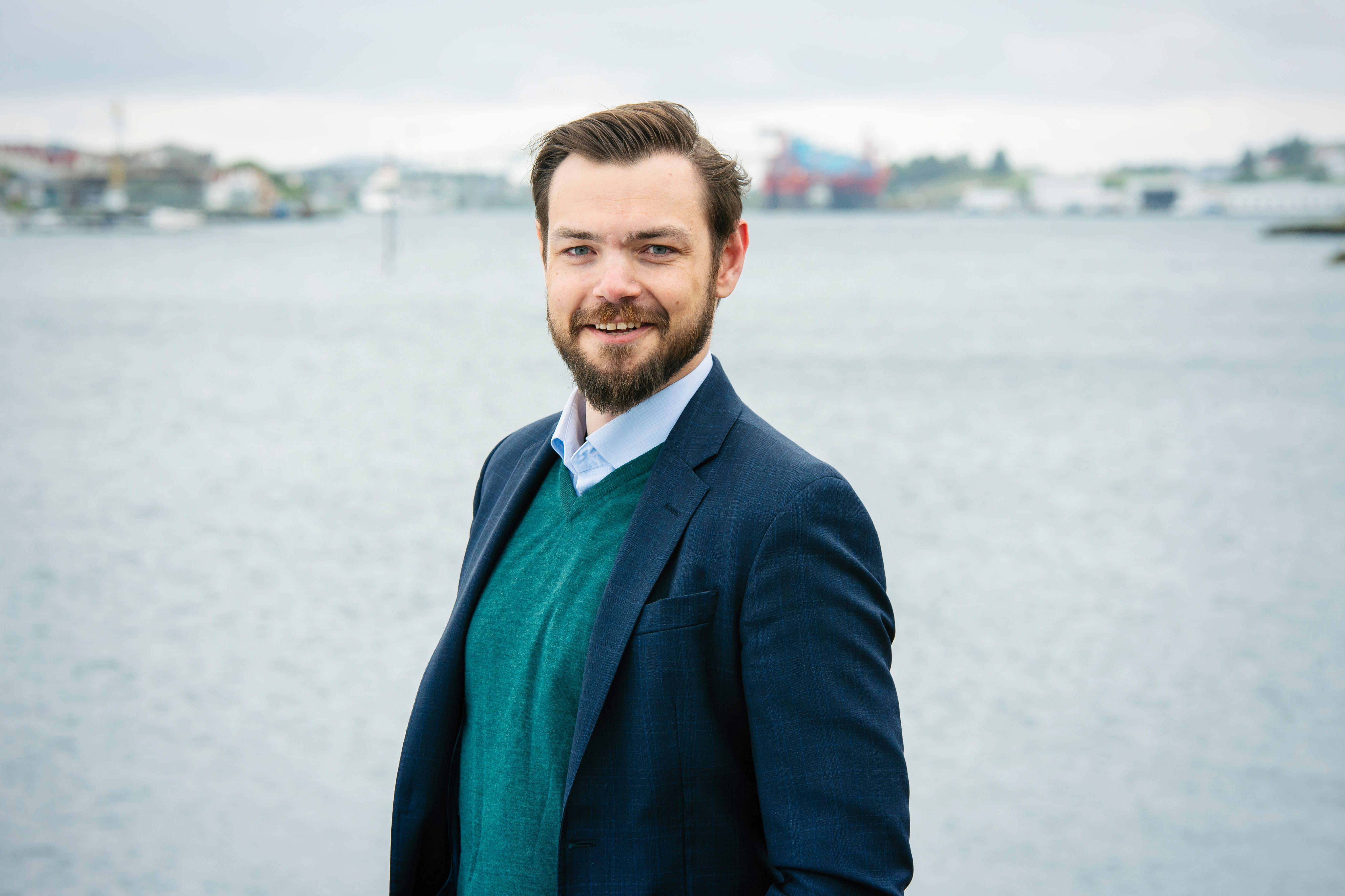 A smiling man with a beard wearing a dark suit jacket and green sweater stands by the water.