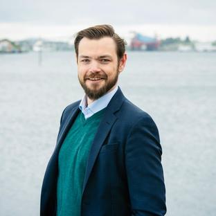 A smiling man with a beard wearing a dark suit jacket and green sweater stands by the water.