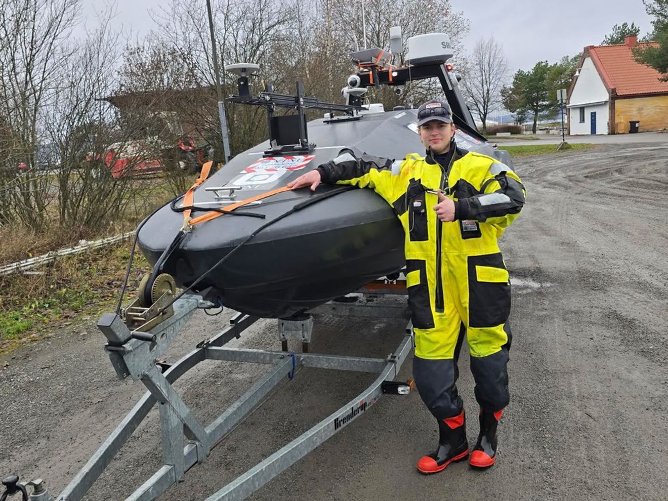 Man in a yellow and black dry suit giving a thumbs-up next to a dark grey unmanned surface vessel on a trailer.