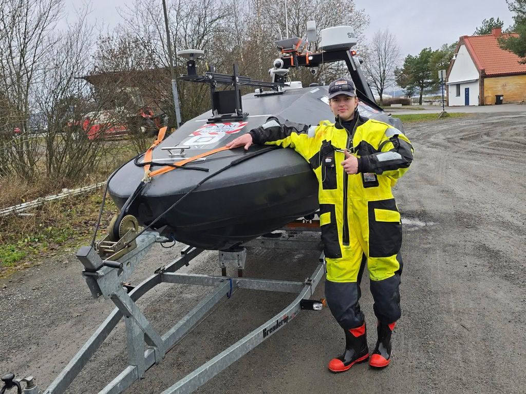 Man in a yellow and black dry suit giving a thumbs-up next to a dark grey unmanned surface vessel on a trailer.