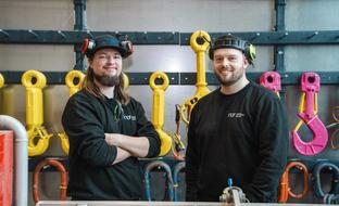 Two smiling men wearing ear protection stand in a facility with industrial lifting hooks and slings.